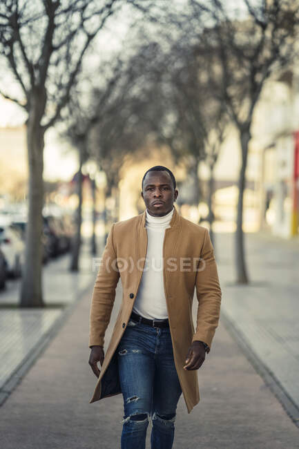 Confiado joven étnico masculino en traje de moda caminando por la calle de la ciudad y mirando a la cámara en el día soleado - foto de stock