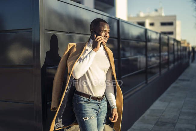Concentrated young African American male in stylish turtleneck and coat talking on mobile phone and looking away while standing on city street — Stock Photo