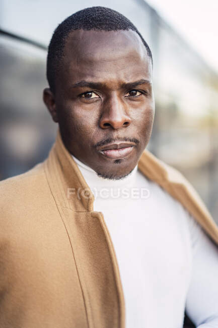 Уверенный молодой афроамериканец в стильном свитере с водолазкой и пальто стоит на улице и смотрит в камеру — стоковое фото