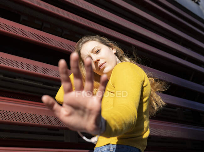 Vista lateral de la joven milenaria pensativa en ropa casual tocando el pelo largo y ondulado y extendiendo la mano hacia la cámara cerca de la cerca de metal rojo - foto de stock