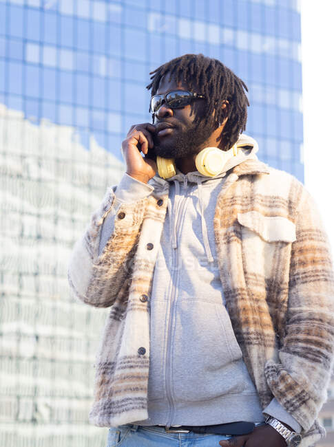 Homme afro-américain à la mode avec des lunettes de soleil et des écouteurs autour du cou parlant sur un téléphone portable tout en se tenant la main dans la poche contre le bâtiment reflétant la ville — Photo de stock