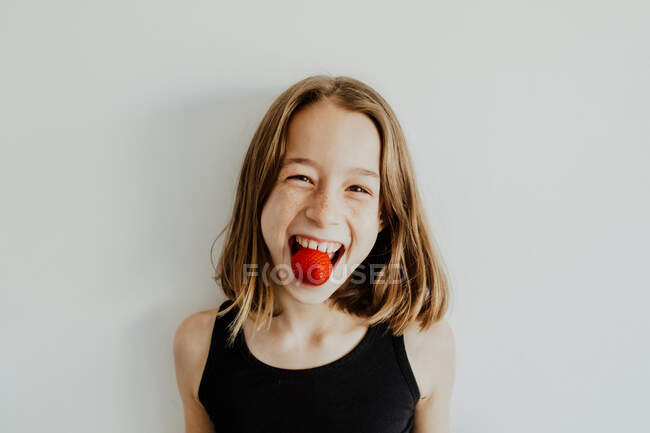 Deleitado adolescente sonriendo y mirando a la cámara mientras muerde sabrosa fresa dulce contra fondo blanco - foto de stock
