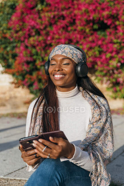 Encantada hembra afroamericana sentada en el parque y escuchando música en auriculares mientras disfruta de canciones con los ojos cerrados - foto de stock