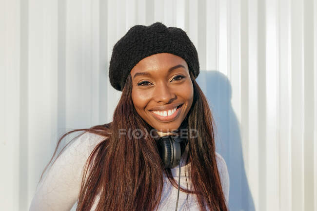 Mujer afroamericana encantada con una sonrisa encantadora y auriculares en el cuello de pie cerca del edificio de la ciudad en el día soleado y mirando a la cámara - foto de stock
