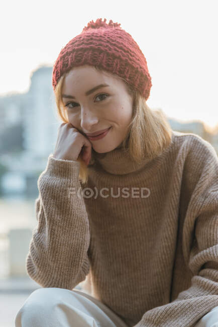 Contenida joven hembra en suéter de punto y sombrero apoyado en la mano y mirando a la cámara mientras está sentada en la calle de la ciudad - foto de stock