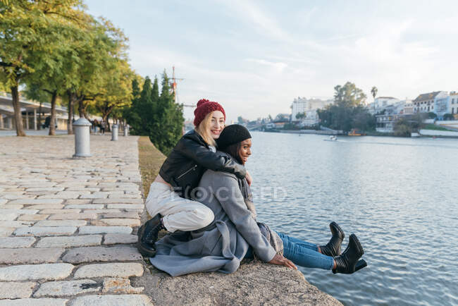 Seitenansicht fröhlicher multiethnischer Freundinnen, die sich auf der Promenade umarmen, während sie das gemeinsame Wochenende im Herbst genießen und wegschauen — Stockfoto