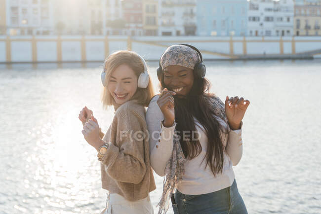 Vista lateral de amigas multirraciales en auriculares disfrutando de la música y bailando juntas en el terraplén en un día soleado - foto de stock