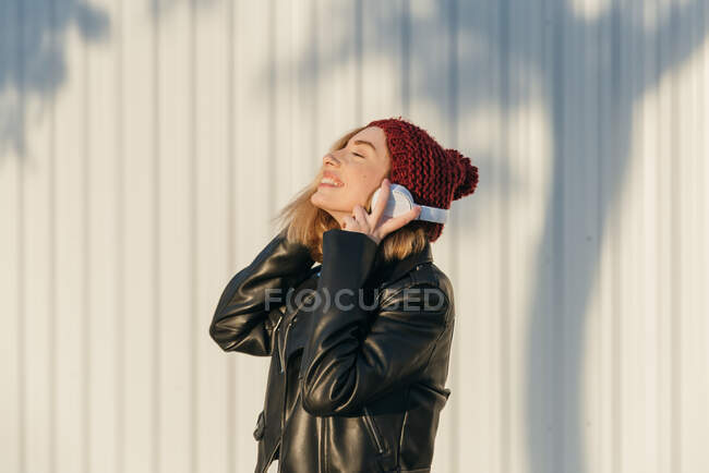 Mulher confiante em roupa de rua e com fone de ouvido em pé na área urbana e olhando para longe — Fotografia de Stock