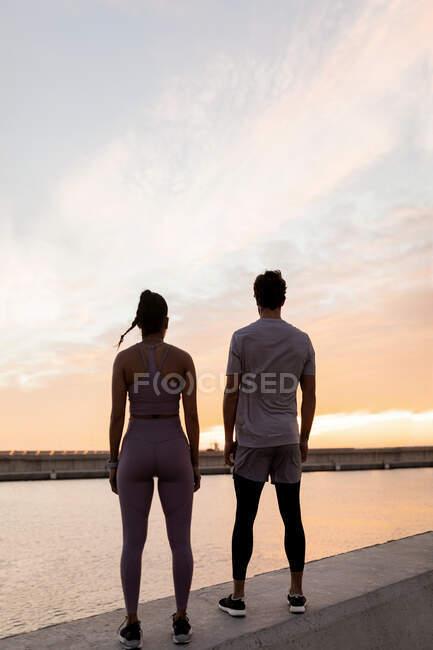 Back view of fit sportswoman with male athlete in sports clothes contemplating lake under cloudy sky at sundown — Stock Photo