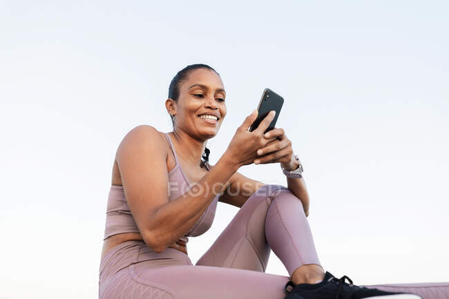 Jeune athlète féminine ethnique en vêtements de sport naviguant sur Internet sur téléphone portable pendant la pause de l'entraînement contre l'océan — Photo de stock