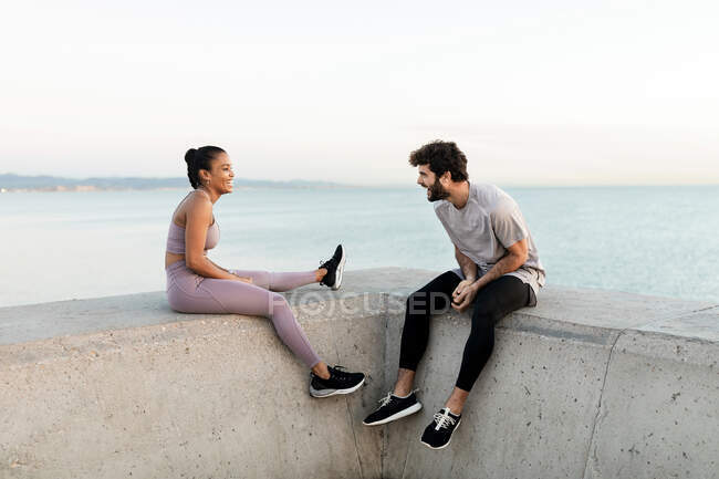 Cheerful young bearded sportsman speaking with content ethnic female athlete while resting against endless sea and looking at each other — Stock Photo