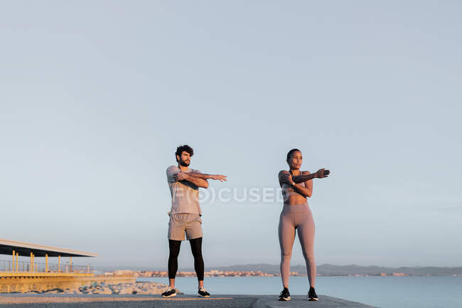 Jóvenes deportistas multirraciales en entrenamiento de ropa deportiva contra el mar mientras miran hacia otro lado bajo el cielo claro - foto de stock