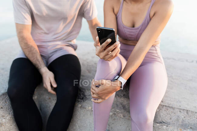 Young glad sportsman with ethnic female partner in sports clothes while spending time together with mobile phone against sea after training — Stock Photo