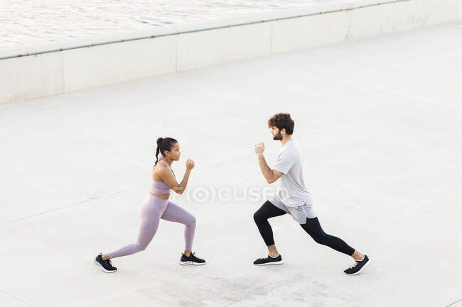 Side view of young determined diverse athletes in sportswear exercising and looking at each other against sea — Stock Photo