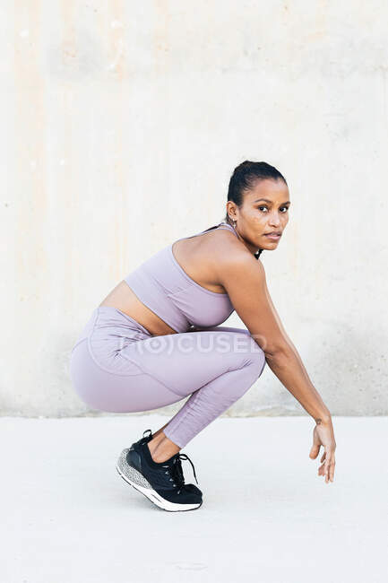 Seitenansicht einer jungen ethnischen Sportlerin in Sportbekleidung und Turnschuhen, die auf Zehenspitzen hockt, während sie auf hellem Hintergrund in die Kamera blickt — Stockfoto