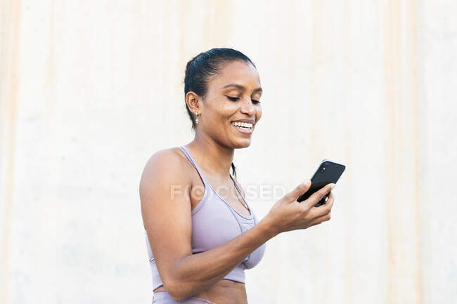 Junge fröhliche ethnische Sportlerin in aktiver Kleidung surft tagsüber mit dem Handy im Internet gegen die Wand — Stockfoto