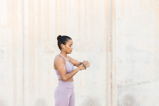 Vista lateral da jovem esportista étnica verificando o tempo enquanto ouve música de fones de ouvido contra a parede áspera — Fotografia de Stock