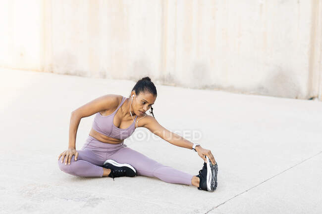 Young fit ethnic female athlete listening to music from earphones while performing forward bend during training on pavement — Stock Photo