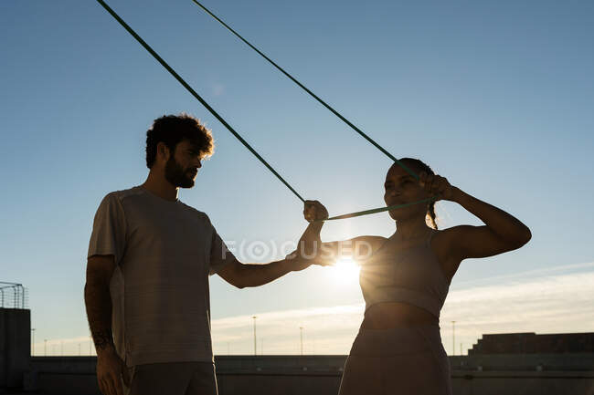 Young bearded sportsman supporting smiling ethnic female athlete during suspension training under blue cloudy sky with shiny sun in back lit — Stock Photo