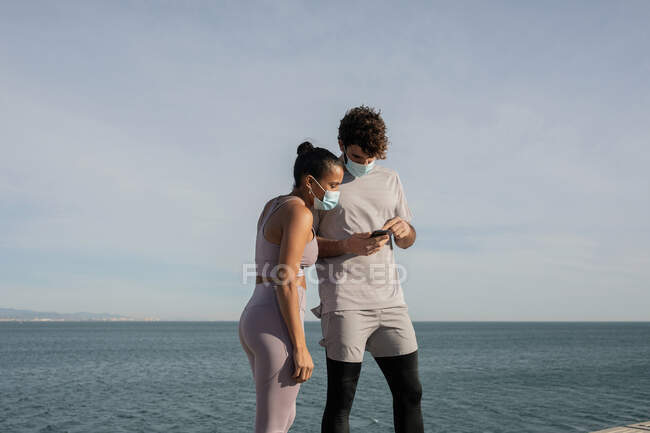Unrecognizable ethnic sportswoman in sterile mask near male partner surfing internet on cellphone against rippled sea under cloudy sky — Stock Photo