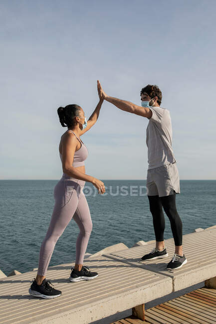 Unerkennbare multirassische Athleten in Sportbekleidung und Gesichtsmasken geben High Five auf einem Damm gegen den Ozean, während sie sich gegenseitig anschauen — Stockfoto