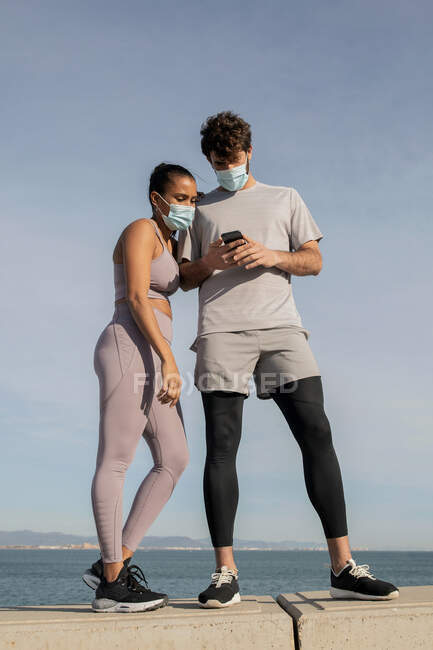 Unerkennbare ethnische Sportlerin mit steriler Maske in der Nähe ihres männlichen Partners, der mit dem Handy gegen das aufgewühlte Meer unter wolkenverhangenem Himmel surft — Stockfoto