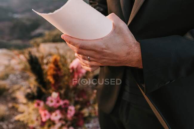 Unrecognizable fiance wearing black suit standing with paper during wedding vow in nature against colorful bouquets of flowers on blurred background — Stock Photo