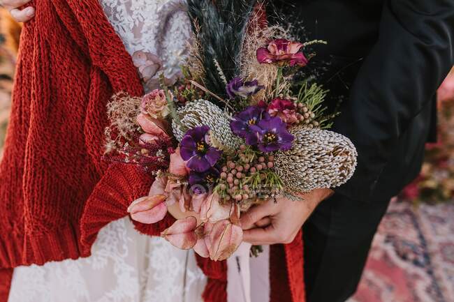 Unrecognizable bride and groom wearing traditional wedding outfits with colorful flowers in hands standing on rug in nature during wedding celebration — Stock Photo