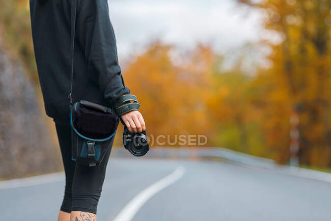 Back view anonymous photographer in casual wear standing with professional photo camera on empty rural road running through yellow forest on autumn day — Stock Photo