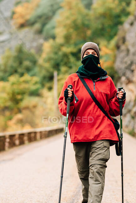 Female hiker in warm clothes with photo camera practicing Nordic walk in autumn forest and looking at camera dreamily — Stock Photo