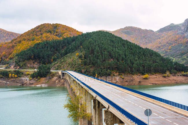 Picturesque scenery of road bridge over blue calm river flowing through forested hills on autumn day — Stock Photo