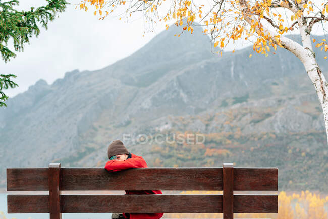 Frau in Oberbekleidung ruht auf Bank im malerischen Herbstpark gegen schwere Bergkette und ruhigen See — Stockfoto