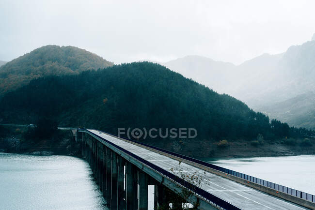 Picturesque scenery of road bridge over blue calm river flowing through forested hills on foggy day — Stock Photo