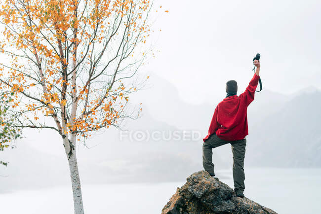 Anonyme Reisende in Oberbekleidung stehen auf massiven Felsen und heben den Arm mit Fotokamera, während sie den nebligen Bergrücken rund um den ruhigen See am Herbsttag bewundern — Stockfoto