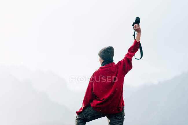 Indietro vista anonimo viaggiatore in capispalla in piedi su roccia massiccia e braccio alzando con macchina fotografica mentre ammirando cresta di montagna nebbioso che circonda lago calmo il giorno d'autunno — Foto stock