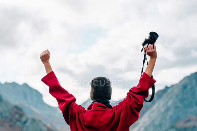 Back view anonymous traveler in outerwear standing on massive rock and raising arm with photo camera while admiring misty mountain ridge surrounding calm lake on autumn day — Stock Photo