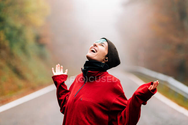 Очарованная молодая женщина в тёплом свитере и шляпе стоит с поднятыми руками и откидывает голову назад, стоя на сельской дороге в туманном осеннем лесу. — стоковое фото