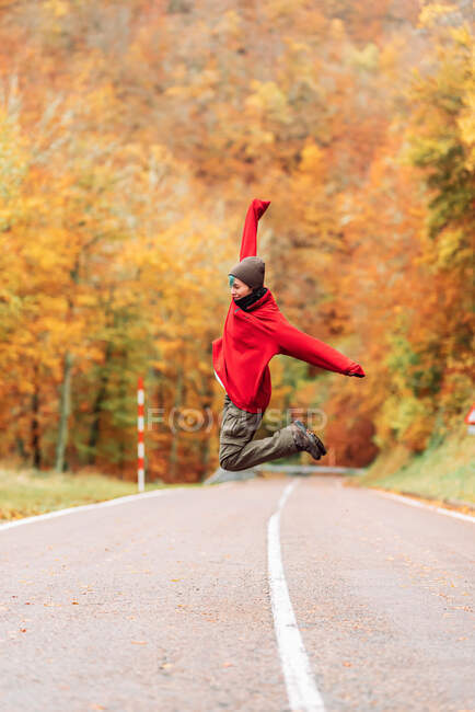 Vue de côté pleine longueur voyageuse insouciante portant des vêtements chauds sautant dans l'excitation au-dessus de la route rurale vide dans la forêt jaune le jour de l'automne — Photo de stock