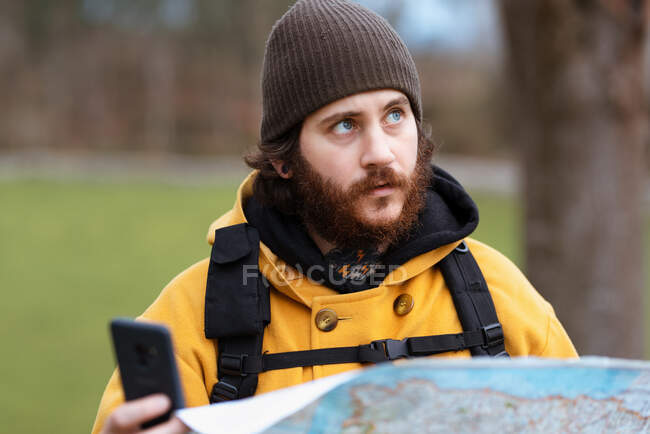 Взрослый созерцательный мужчина путешественник в верхней одежде с мобильным телефоном и бумажной картой, глядя вверх в дневное время — стоковое фото