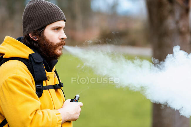 Side view of unshaven male traveler in outerwear with e liquid bottle vaping and looking forward on meadow — Stock Photo