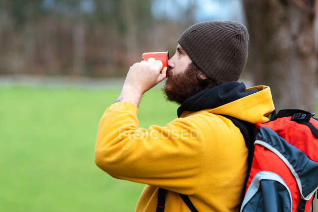 Side view of bearded male trekker with rucksack drinking tea from metal mug on meadow — Stock Photo
