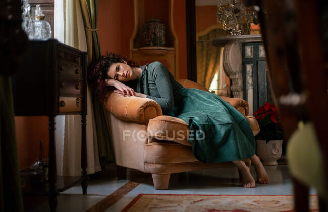 Ganzkörper barfuß elegante Frau auf Sessel blickt in die Kamera, während sie sich im Zimmer mit Spiegel und Vintage-Interieur ausruht — Stockfoto