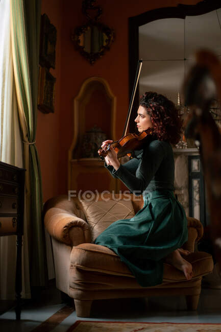 Vue latérale complète du corps d'une musicienne qualifiée jouant du violon alors qu'elle était assise sur un fauteuil dans une salle de style vintage pendant la répétition — Photo de stock