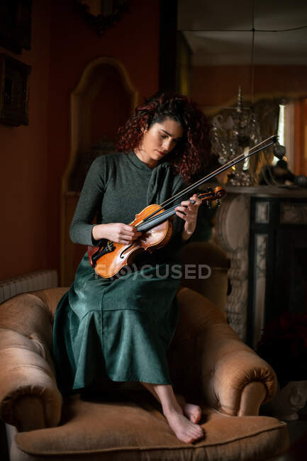 Músico femenino sosteniendo violín mientras está sentado en un sillón en una habitación de estilo vintage durante el ensayo - foto de stock