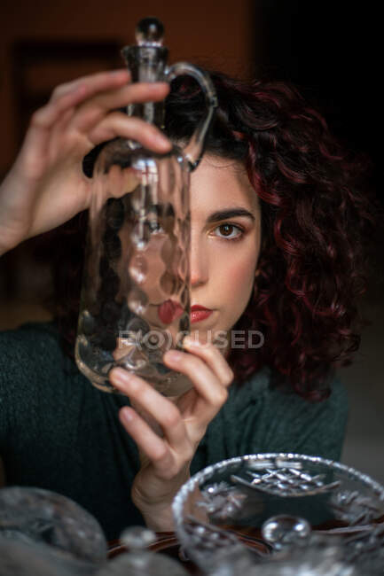 Aufmerksame Frau mit schwarzem lockigem Haar blickt durch transparente Glaskanne, während sie in der Nähe von Vintage-Kristallgläsern im Zimmer steht — Stockfoto