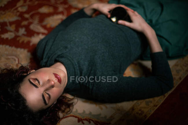 Von oben sieht man eine entspannte Frau mit schwarzen Haaren, die mit geschlossenen Augen auf einem Teppich liegt und eine transparente Kristallkugel im Raum mit Retro-Design hält — Stockfoto