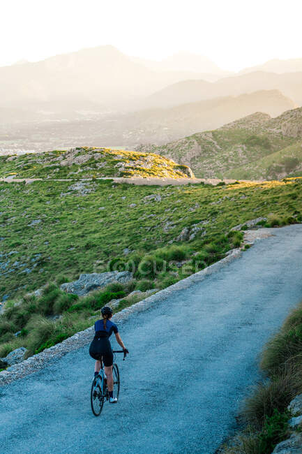 Vue de dos de cycliste anonyme en tenue décontractée à vélo seul dans les hautes terres verdoyantes avec des montagnes majestueuses dans la brume — Photo de stock