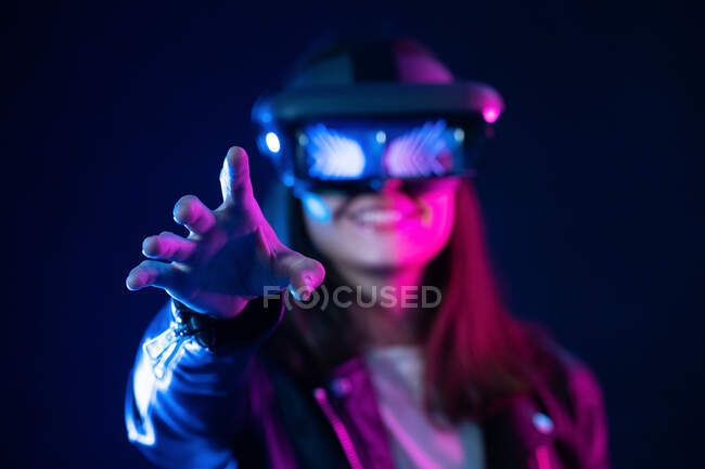 Неузнаваемая женщина с вытянутой рукой в VR гарнитуре, исследуя виртуальную реальность под синим неоновым светом — стоковое фото