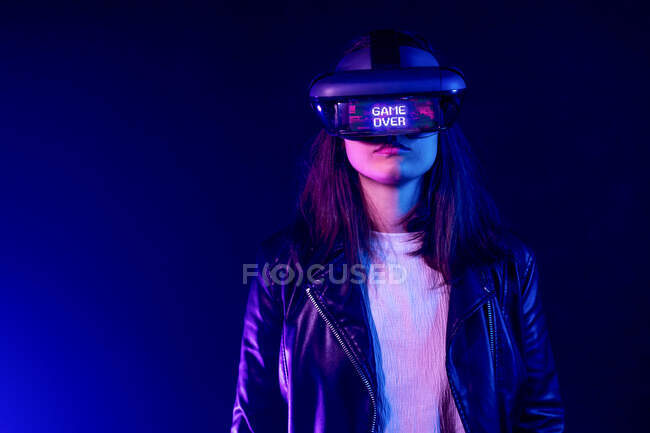 Unrecognizable female wearing modern headset with Game Over inscription while exploring virtual reality in dark room with neon light near wall — Stock Photo