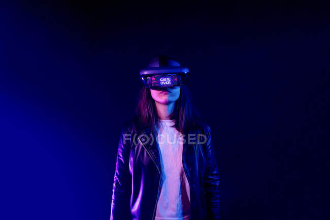 Unerkennbare Frau trägt modernes Headset mit Game Over-Aufschrift, während sie die virtuelle Realität im dunklen Raum mit Neonlicht in Wandnähe erkundet — Stockfoto
